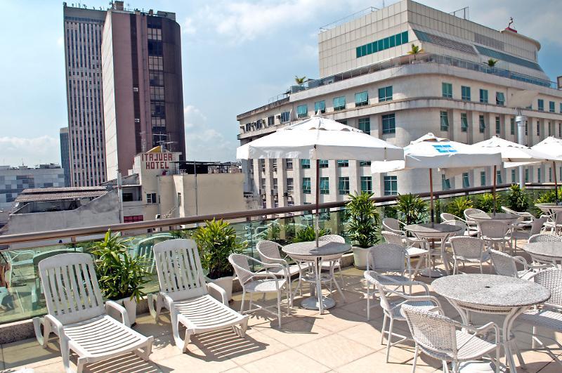 Hotel Atlântico Business Centro Rio de Janeiro Exterior foto The photo shows a rooftop area with several white lounge chairs and circular tables. There are umbrellas providing shade over some of the seating. In the background, there are tall buildings, including a modern skyscraper with a distinct architectura